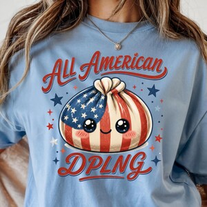 May include: Light blue t-shirt with the words "All American DPLNG" in red script. The graphic features a dumpling with an American flag design and cute eyes, surrounded by stars.