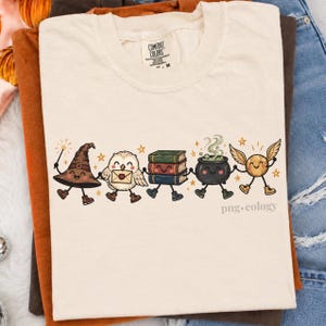 May include: A cream-colored t-shirt featuring a graphic of five anthropomorphic characters holding hands: a brown wizard hat, a white owl with a letter, a stack of three books, a black cauldron with green steam, and a golden winged ball.
