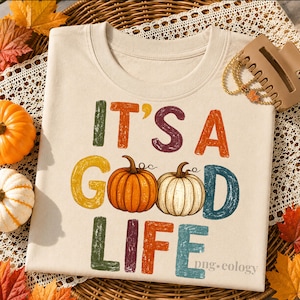 Puede incluir: Sudadera crema con la frase "IT'S A GOOD LIFE" en letras coloridas y texturizadas. El diseño presenta ilustraciones de dos calabazas. La sudadera está rodeada de decoraciones otoñales, incluyendo calabazas y hojas.