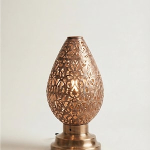 Peut inclure: Une lampe de table en forme de goutte d'eau avec une finition bronze métallique. Le corps présente un motif floral complexe découpé au laser qui laisse passer la lumière. Elle repose sur une base circulaire en bronze avec un cordon d'alimentation.