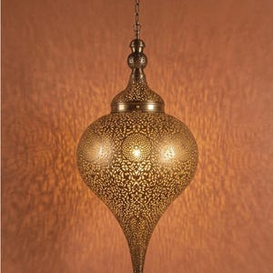 Op de afbeelding: Een druppelvormige hanglamp van antiek messingkleurig metaal met ingewikkelde, lasergesneden filigraanpatronen. De lamp hangt aan een bijpassende ketting en plafondkap en werpt een warme, gedessineerde gloed op de muur erachter.
