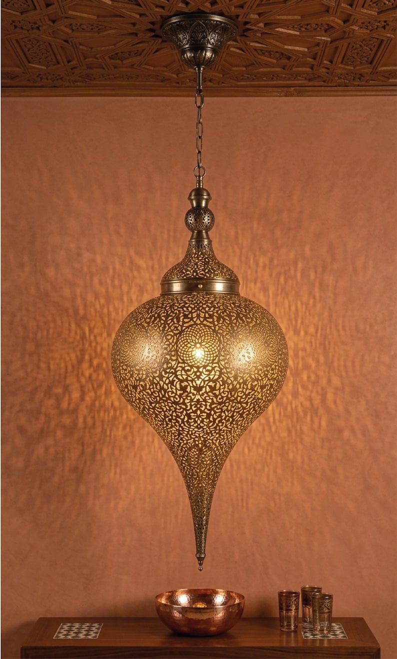 Op de afbeelding: Een druppelvormige hanglamp van antiek messingkleurig metaal met ingewikkelde, lasergesneden filigraanpatronen. De lamp hangt aan een bijpassende ketting en plafondkap en werpt een warme, gedessineerde gloed op de muur erachter.