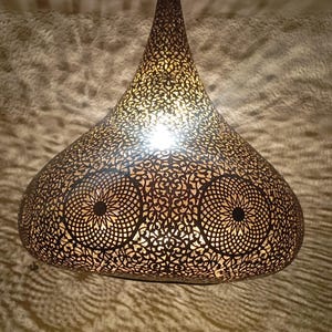 Puede incluir: Una lámpara colgante decorativa con un intrincado diseño calado. La lámpara tiene forma de lágrima, con una parte superior puntiaguda y una base más ancha y redondeada. El metal es de un color oscuro y cálido, y la luz brilla a través de los patrones recortados.