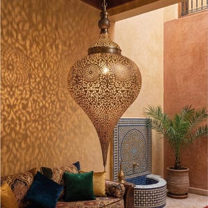 Op de afbeelding: Een grote, druppelvormige hanglamp van geperforeerd messing met ingewikkelde, herhalende geometrische patronen. Het metalen oppervlak heeft een warme afwerking die gedetailleerde, sierlijke schaduwen op de omliggende muren werpt.