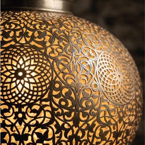 Op de afbeelding: Een bolvormige metalen hanglamp met een ingewikkeld, lasergesneden filigraanpatroon van herhalende florale en geometrische motieven. Het donkere, antiek afgewerkte metalen oppervlak contrasteert met het warme, gouden licht van binnenuit.