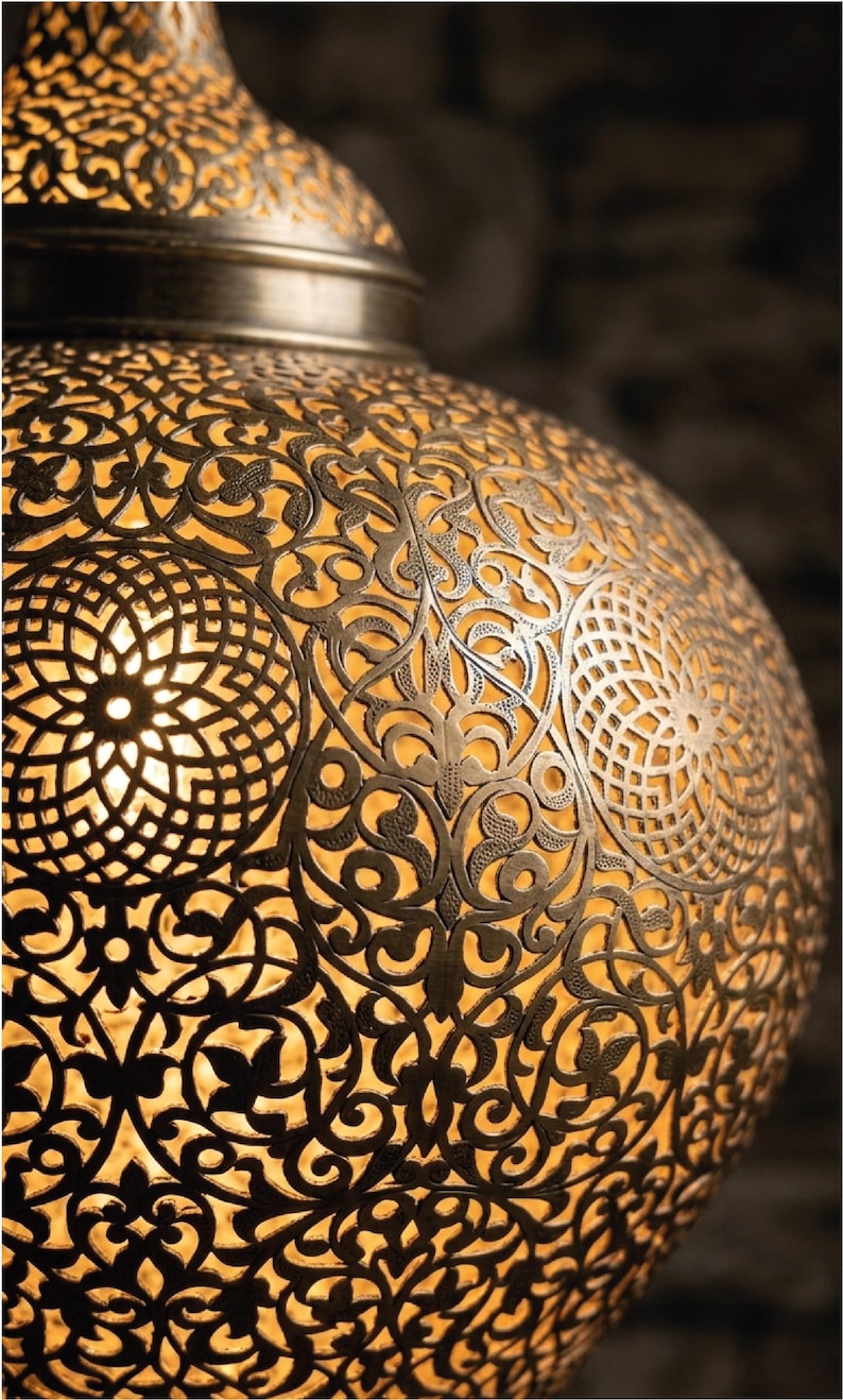 Op de afbeelding: Een bolvormige metalen hanglamp met een ingewikkeld, lasergesneden filigraanpatroon van herhalende florale en geometrische motieven. Het donkere, antiek afgewerkte metalen oppervlak contrasteert met het warme, gouden licht van binnenuit.