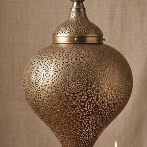 May include: A teardrop-shaped hanging pendant lamp made of antique brass-colored metal with intricate laser-cut geometric patterns. The lamp features a pointed base and a matching decorative finial at the top, suspended by a metal chain.