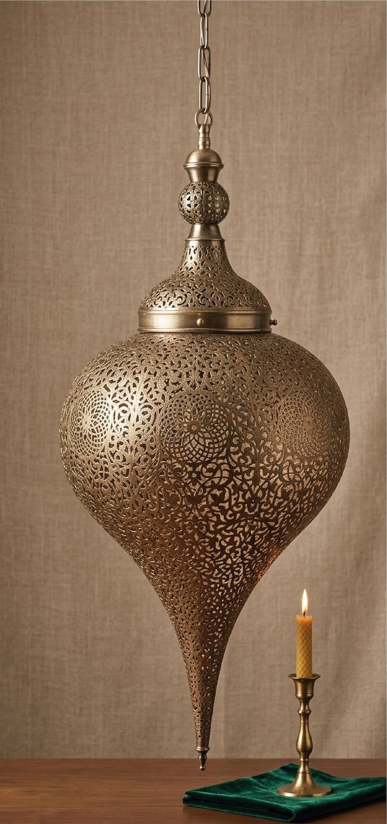 Op de afbeelding: Een druppelvormige hanglamp van antiek messingkleurig metaal met ingewikkelde, lasergesneden geometrische patronen. De lamp heeft een puntige basis en een bijpassende decoratieve knop aan de bovenkant, opgehangen aan een metalen ketting.