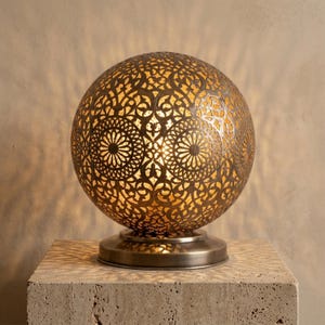 Peut inclure: Une lampe de table sphérique en métal avec un motif en filigrane découpé au laser. La surface couleur bronze présente des motifs circulaires et floraux répétitifs qui laissent passer une lumière chaude à travers les découpes.