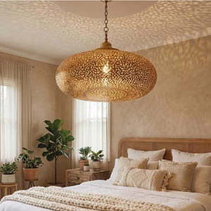 May include: A large, round, gold-toned metal pendant light fixture with an intricate, laser-cut filigree pattern. The lamp is suspended from the ceiling by a chain and casts a detailed, lace-like shadow pattern across the ceiling surface.