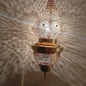 May include: A hanging bronze-colored pendant lamp with an intricate, openwork design. The lamp has a teardrop shape with a decorative pattern that casts patterned shadows on the wall. The lamp is lit from within, creating a warm glow.