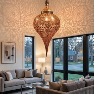 Op de afbeelding: Een grote, druppelvormige hanglamp gemaakt van metaalachtig bronskleurig materiaal met ingewikkelde, lasergesneden bloemmotieven. De lamp hangt aan een dunne ketting en werpt gedetailleerde, uitgestrekte schaduwpatronen op de omliggende muren.