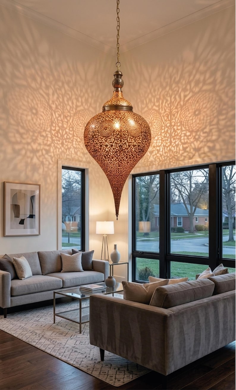 Op de afbeelding: Een grote, druppelvormige hanglamp gemaakt van metaalachtig bronskleurig materiaal met ingewikkelde, lasergesneden bloemmotieven. De lamp hangt aan een dunne ketting en werpt gedetailleerde, uitgestrekte schaduwpatronen op de omliggende muren.