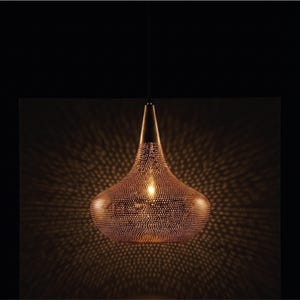 Peut inclure: Une suspension en forme de goutte d'eau en métal de couleur bronze, dotée d'un motif dense de petites perforations circulaires. La lumière diffuse un éclat chaud à travers les trous, projetant un motif d'ombre radial sur le fond sombre.