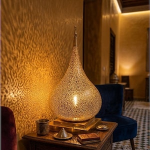 May include: A teardrop-shaped brass table lamp with intricate laser-cut perforations that cast a detailed light pattern on the wall. The lamp sits on a square brass base and features a pointed finial at the top, emitting a warm golden glow.