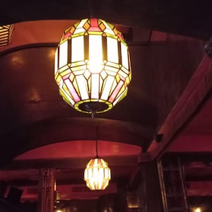 May include: Two ornate, hanging pendant lamps with geometric stained glass designs. The lamps are illuminated, casting a warm glow in a dimly lit room. The lampshades feature a mix of yellow, pink, and white colors. The room has a dark, reddish-brown color scheme.