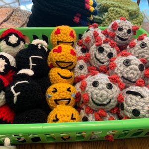 May include: A green plastic basket filled with various crocheted items. There are several emoji-faced balls in yellow, black, and white, along with red and white balls. Also, there are gray crocheted balls with red accents and button eyes.