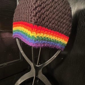 May include: A dark brown crocheted beanie with a vibrant rainbow band at the base. The rainbow features stripes of red, orange, yellow, green, blue, and purple. The hat is displayed on a black wig stand.
