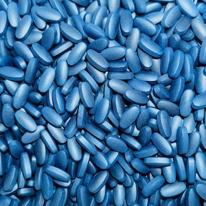 May include: A close-up shot of a large pile of blue oval-shaped pills. The pills are uniformly colored and densely packed, creating a textured surface. The lighting highlights the rounded edges and smooth surfaces of the pills.