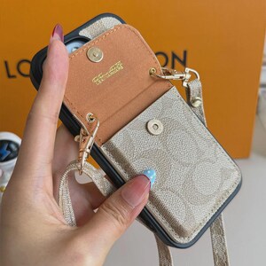 以下が含まれることがあります： ベージュとタンの柄入りスマートフォンケース。スナップボタン付きの取り外し可能なポーチが付いています。ポーチの内側はタンカラーで、ゴールドで「LEATHERWARE」の文字がプリントされています。同色のベージュのストラップが金属製クリップで取り付けられています。