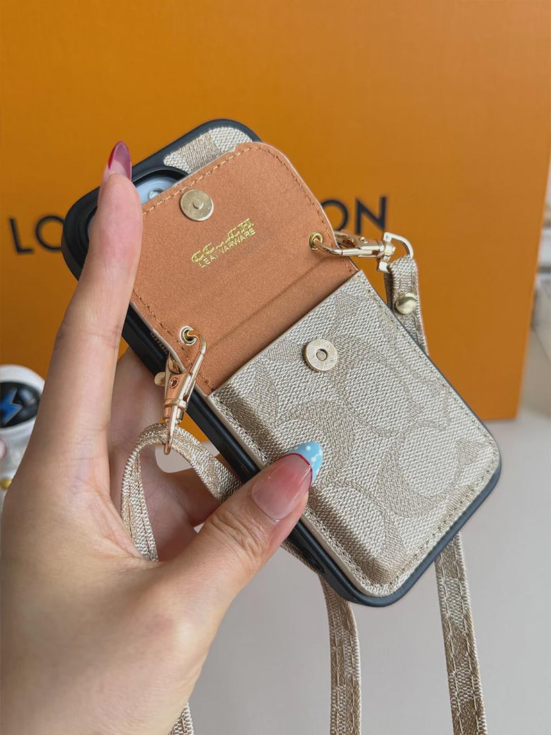 以下が含まれることがあります： ベージュとタンの柄入りスマートフォンケース。スナップボタン付きの取り外し可能なポーチが付いています。ポーチの内側はタンカラーで、ゴールドで「LEATHERWARE」の文字がプリントされています。同色のベージュのストラップが金属製クリップで取り付けられています。