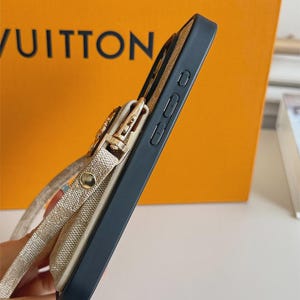 以下が含まれることがあります： 黒いスマートフォンケースで、側面にベージュの柄入りポーチが付いています。ゴールドトーンの金属製クラスプと、お揃いのベージュのストラップが特徴です。ケースには側面のボタンが見え、デバイスをしっかりと保持しつつ収納できるように設計されたスリムなプロファイルです。
