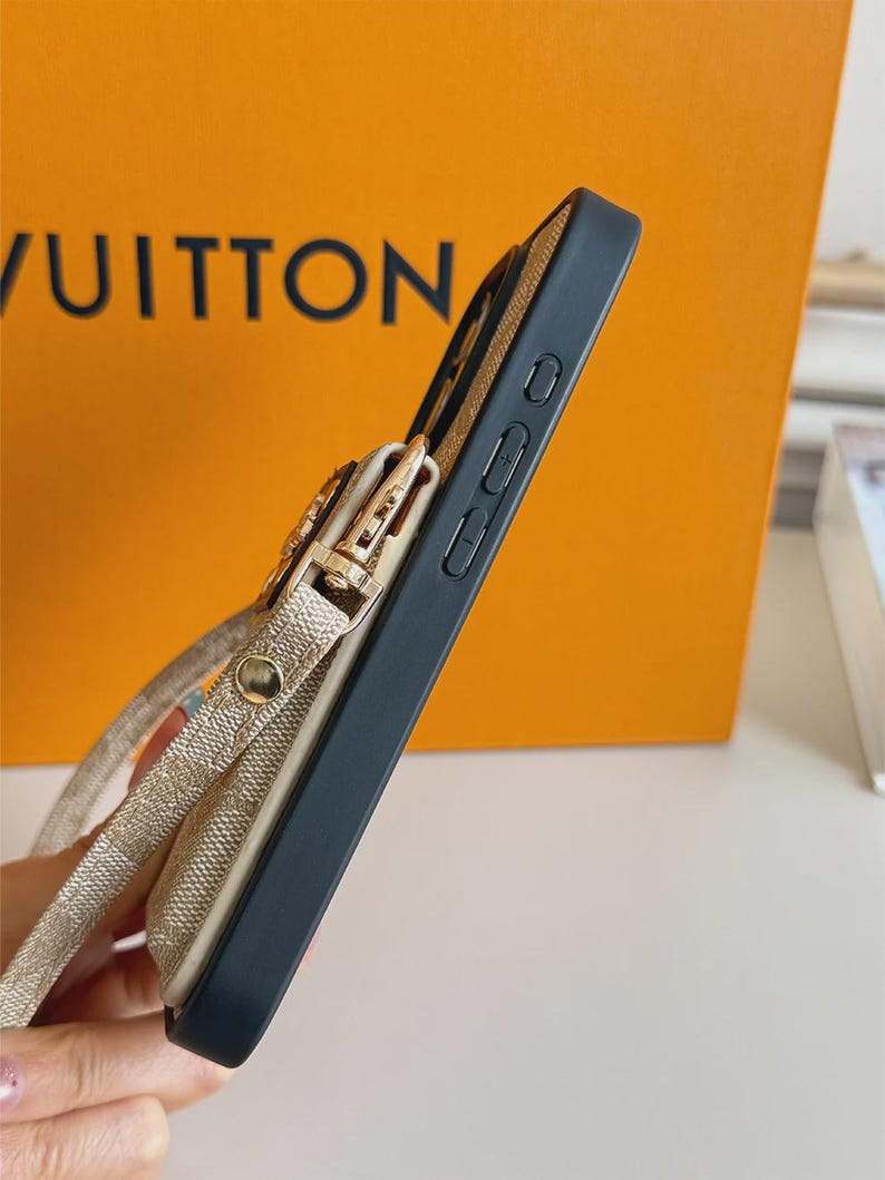 以下が含まれることがあります： 黒いスマートフォンケースで、側面にベージュの柄入りポーチが付いています。ゴールドトーンの金属製クラスプと、お揃いのベージュのストラップが特徴です。ケースには側面のボタンが見え、デバイスをしっかりと保持しつつ収納できるように設計されたスリムなプロファイルです。