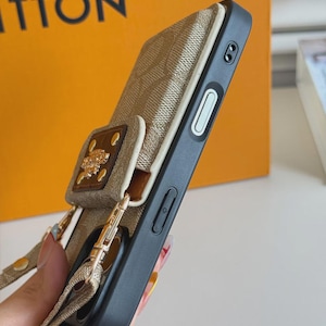 以下が含まれることがあります： 繰り返しモノグラム柄が施されたベージュのスマートフォンケース。ゴールドトーンのメタルエンブレムと4つのゴールドリベットが付いた取り外し可能なカードホルダーが特徴です。黒いフレーム、サイドボタン、同色のベージュの布製リストストラップが付いています。