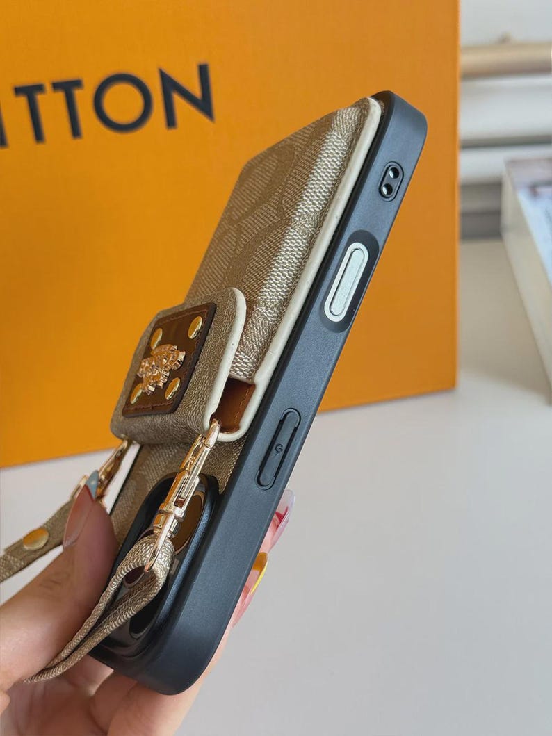 以下が含まれることがあります： 繰り返しモノグラム柄が施されたベージュのスマートフォンケース。ゴールドトーンのメタルエンブレムと4つのゴールドリベットが付いた取り外し可能なカードホルダーが特徴です。黒いフレーム、サイドボタン、同色のベージュの布製リストストラップが付いています。
