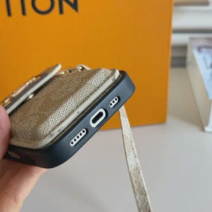 以下が含まれることがあります： モノグラム柄のベージュの背面アタッチメントと、同色のベージュの布製ストラップが付いた黒いスマートフォンケース。ケースの底面には、中央の充電ポート用カットアウトと、その両側に丸い穴が開いた2つのスピーカーグリルがあります。