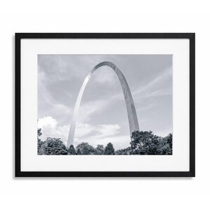 May include: Black and white framed print featuring the Gateway Arch in St. Louis, Missouri. The arch is a prominent, curved structure set against a cloudy sky, with trees visible at the base. The print is encased in a black frame.