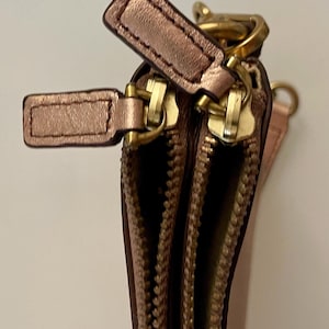 May include: Close-up of a rose gold leather wallet with two zippered compartments. The wallet features gold-colored zippers and pull tabs. The leather has a slight sheen, and the stitching is visible.