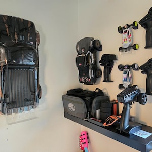 Può includere: Una collezione di veicoli radiocomandati e accessori montati su una parete e una mensola. L'esposizione include un grande camion nero, due buggy più piccoli, tre trasmettitori neri a impugnatura a pistola, una borsa grigia e pneumatici.