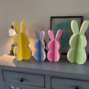 May include: Four wooden Easter bunny decorations in yellow, blue, pink, and green. Each bunny has a layered design with a painted front and natural wood edges. They are displayed on a gray table.