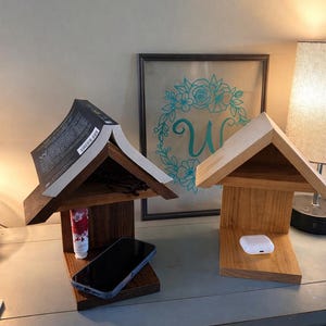 Puede incluir: Dos organizadores de mesita de noche de madera con forma de casa para pájaros. Uno contiene un libro, gafas y un teléfono; el otro contiene un par de auriculares inalámbricos blancos. Los organizadores son de madera marrón clara y oscura.