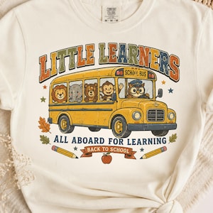 Op de afbeelding: Een crèmekleurig T-shirt met korte mouwen en een afbeelding van een gele schoolbus met een leeuw, olifant, beer, aap en uil. De tekst luidt LITTLE LEARNERS, ALL ABOARD FOR LEARNING en BACK TO SCHOOL, met potloden en een appel.