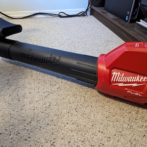 May include: A red and black Milwaukee FUEL cordless leaf blower. The blower has a curved nozzle and the Milwaukee logo is prominently displayed on the side. The blower is designed for outdoor use.