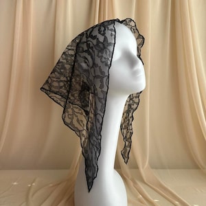 May include: A black lace head covering with a delicate floral pattern and scalloped edges, draped over a white mannequin head. The fabric is sheer and lightweight, featuring an intricate openwork design that creates a detailed texture.