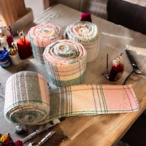 May include: Several rolls of plaid fabric in shades of pink, green, and cream are displayed on a wooden surface. One roll is unraveled, showing the pattern. The scene includes crafting tools and supplies, suggesting a textile project.