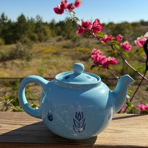 May include: A light blue ceramic teapot with a matching lid, featuring a dark blue tulip illustration on the front and smaller tulip motifs around the body. The teapot has a curved handle and a short spout, with a smooth, glossy finish.