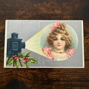May include: A vintage Christmas postcard featuring a blue magic lantern projecting a circular image of a young person with curly hair, a pink bow, and a pearl necklace. The text reads Best Wishes for Christmas, accompanied by green holly leaves and red berries.