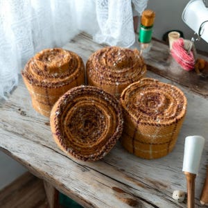 May include: Four rolled fabric bundles in shades of brown and tan with a plaid pattern. The bundles are arranged on a weathered wooden surface. The fabric appears to be a textured material, possibly wool or a similar textile.