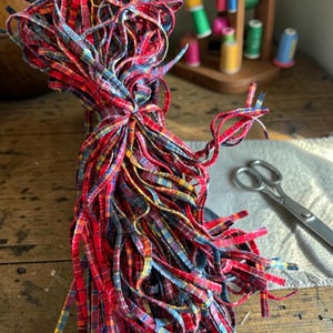 May include: A bundle of thin fabric strips featuring a red, blue, and yellow plaid pattern. The strips are gathered together in the center, creating a cascading effect with loose ends hanging down in various directions, showcasing the textile texture.