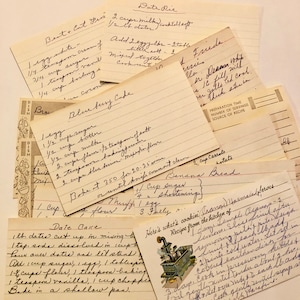 May include: A collection of vintage recipe cards with handwritten instructions for baking blueberry cake, banana bread, date pie, and date cake. The cards are yellowed with age and have a rustic, charming aesthetic.