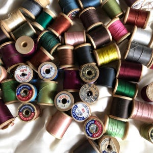 May include: A collection of vintage wooden spools of thread in various colors, including green, pink, brown, and blue. Some spools have labels with text like "COATS & CLARKS" and "MERCE." The spools are scattered on a white surface.