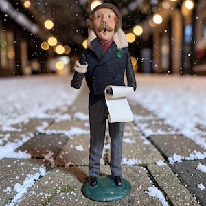 May include: A figurine of a person with a brown mustache wearing a brown cap, a dark gray double-breasted coat with a cream-colored collar, and gray pinstriped trousers. The figure holds a blank white scroll and stands on a round green base.