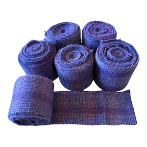 May include: Six rolls of blue and purple plaid fabric, with one roll unraveled. The fabric has a woven texture and a pattern of intersecting lines. The rolls are tightly wound, and the unraveled piece lies flat.