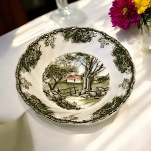 May include: A white ceramic plate with a scalloped edge and a green vine pattern. The center of the plate features a pastoral scene with a house, trees, and a fence. The plate is likely vintage or antique.