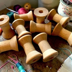 May include: A collection of light brown wooden spools scattered with colorful thread remnants, including dark green, red, and purple strands, alongside a metal sewing needle and a small wooden bead on a dark surface.