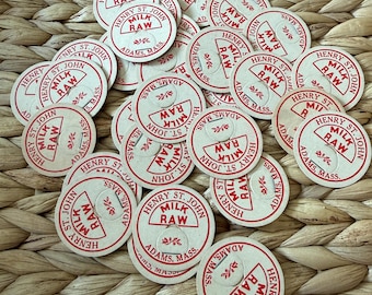 May include: A collection of circular off-white paper milk bottle caps featuring red printed text that reads Henry St. John, Milk Raw, and Adams, Mass. Each cap includes a small tab and a decorative floral graphic element.